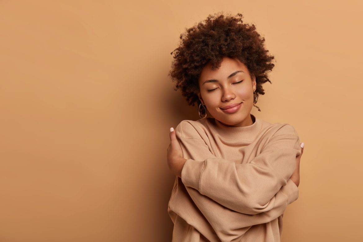 love yourself, self acceptance. gentle lovely afro american woman crosses hands and embraces own body, tilts head and closes eyes, wears comforable brown sweater, models indoor. i love myself